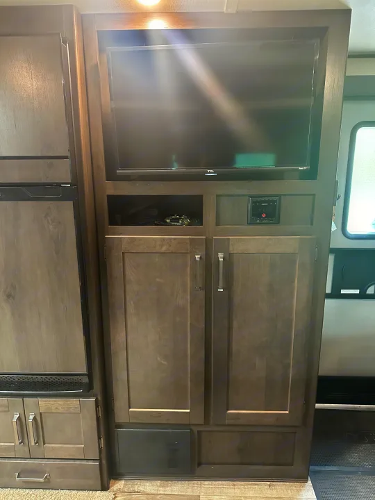 Modern RV rental interior: a fridge on the left, with an entertainment center featuring a TV, Furrion stereo, and cabinets.. Grand Design Transcend Xplor 2022