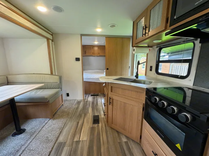 Bright RV rental interior: kitchen with sink & oven, dining booth, and peek into the bedroom. Modern, light wood finishes.. Prime Time Avenger 2022