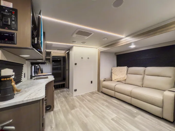 Modern RV rental interior, featuring a bright, spacious living area with a reclining sofa, a compact kitchen, and overhead lighting.. Grand Design Transcend Xplor 2024
