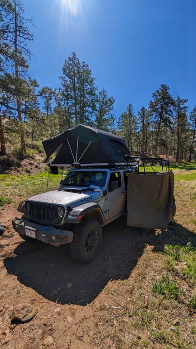 Jeep RV rental with rooftop tent & privacy screen set up for overlanding and camping in a sunny forest.. Jeep Wrangler 2024