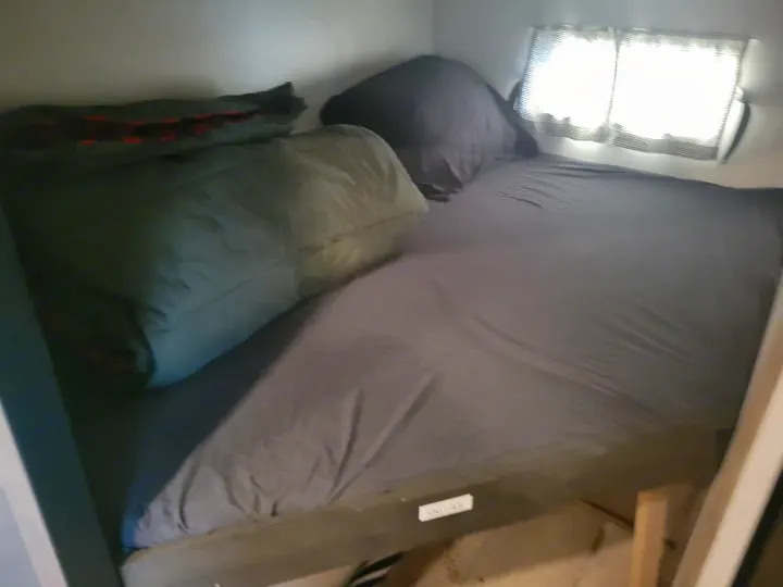 RV rental interior: A bed with grey sheets, pillows, and a sleeping bag, lit by a window with striped blinds.. KZ Confluence 2022