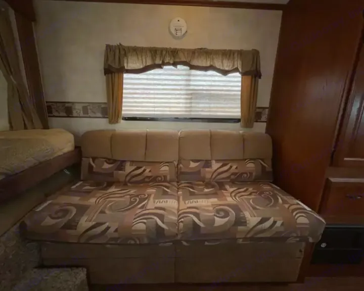 RV rental interior: patterned sofa, window with blinds, and a visible bed. Wood accents add warmth to this mobile living space.. Keystone RV Outback 2010