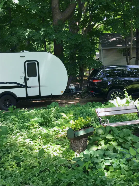 White camper (17 BH) and black SUV, an RV rental setup, parked amidst dense green trees and plants.. Braxton Creek Bushwhacker Plus 2021