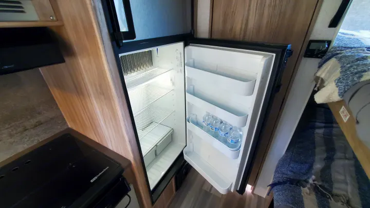 Inside an RV rental: a compact refrigerator with shelves and water bottles, next to a stove and bed.. Winnebago Minnie Winnie 2021