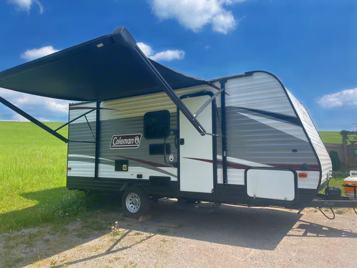 Experience camping with this Coleman RV rental. Grey & white travel trailer with extended black awning on gravel.. Coleman 17FQ 2019