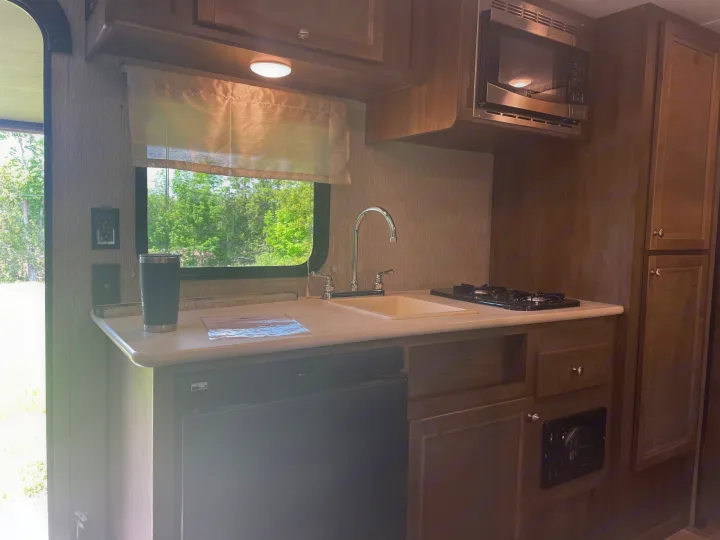 RV rental kitchen interior: microwave above counter with sink, stove, and an under-counter fridge next to a window.. Coleman 17FQ 2019