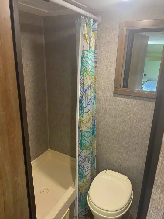 Compact rv rental bathroom featuring a corner shower with a tropical leaf curtain, a toilet, and a mirror.. Coleman 17FQ 2019