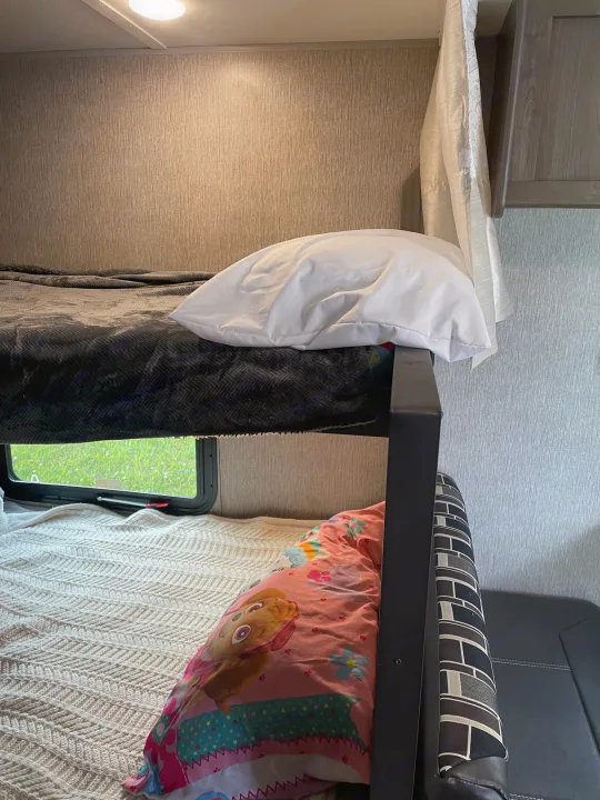 RV rental interior: stacked bunk beds. Upper has grey blanket, lower a knitted blanket with character pillow. Window view of grass.. Coleman 17FQ 2019
