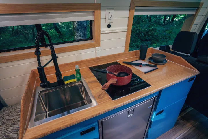 Compact RV rental kitchen with bamboo counters, sink, induction cooktop, and fridge, offering forest views.. Mercedes-Benz Sprinter 2024