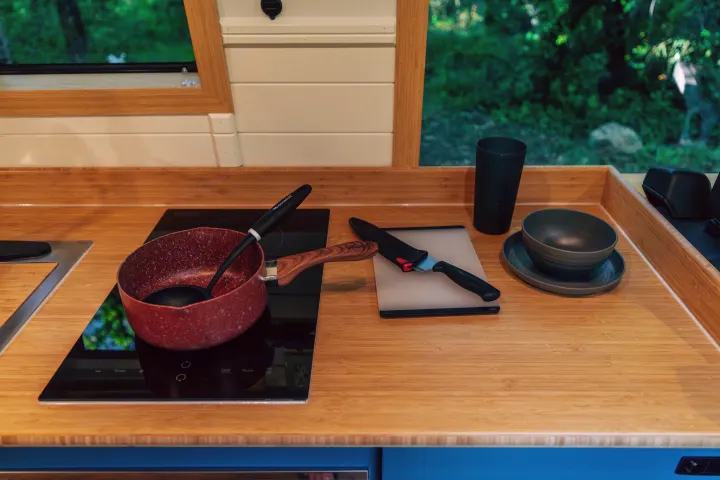 An RV rental's compact kitchen. Bamboo counter, induction cooktop, pot, knife, and dishes by a window overlooking trees.. Mercedes-Benz Sprinter 2024