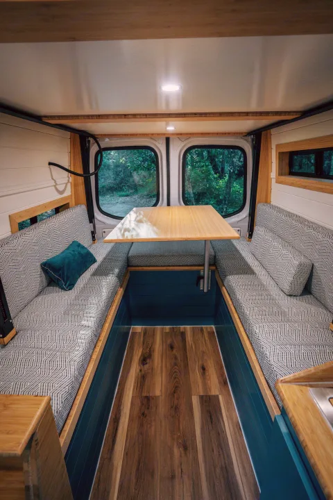 Cozy rv rental interior: wood-paneled van dining area with patterned cushions, overhead light, and forest views.. Mercedes-Benz Sprinter 2024