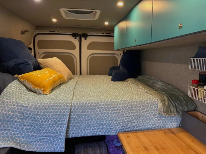 Cozy RV rental interior: a comfy bed with blue bedding, teal overhead cabinets, and compact storage for travel.. RAM ProMaster 2023