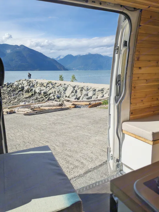 RV rental view from inside a campervan: snowy mountains, inlet, and two people on a rocky shore. Great for travel.. Dodge Sprinter Van 2007