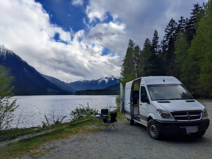 Scenic RV rental: White camper van by a lake, chairs out, facing snow-capped mountains and cloudy skies.. Dodge Sprinter Van 2007