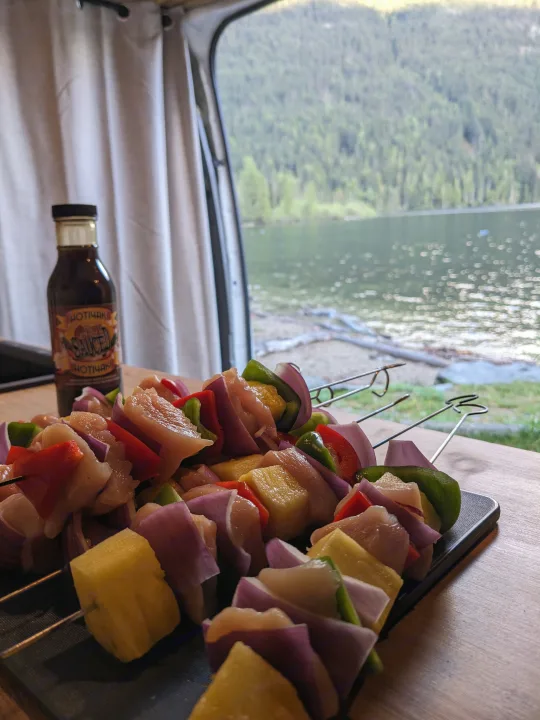 RV rental meal prep: chicken, pineapple & veggie skewers with sauce, set against a scenic lake & forest view. Camping dinner.. Dodge Sprinter Van 2007