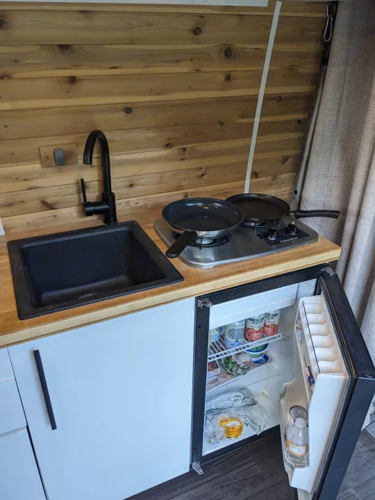 Compact RV rental kitchen with wood walls, a black sink, two-burner Suburban stove, and an open, stocked mini-fridge.. Dodge Sprinter Van 2007