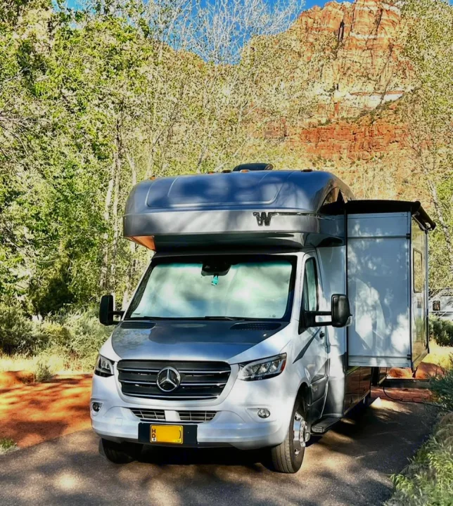Silver Mercedes RV rental parked amidst green trees with a stunning red rock mountain backdrop. Ideal for nature adventures.. Winnebago View 2022