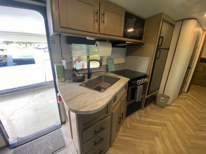 Inside a modern **rv rental** kitchen featuring a sink, microwave, and stove. The open door offers a view of the campground.. Cruiser RV Shadow Cruiser 2021