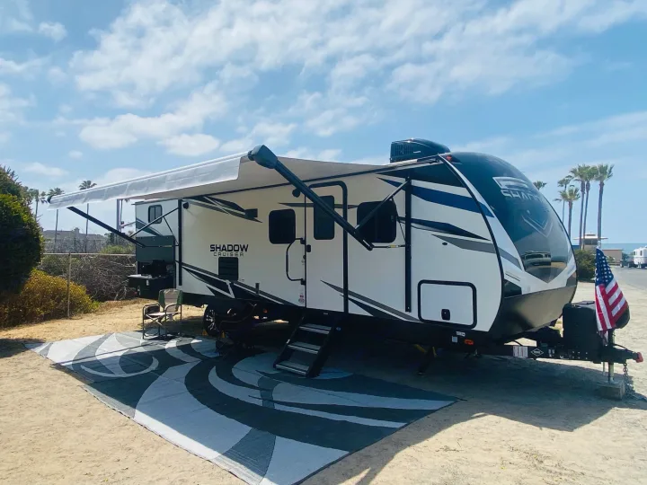 Coastal RV rental campsite features a white travel trailer, extended awning, ground mat, and US flag.. Cruiser RV Shadow Cruiser 2021