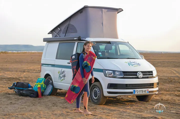 Kiteboarding adventure: A woman with her board stands by a white RV rental camper van with pop-up roof on a sandy beach.. Volkswagen California Coast T6.1 DSG7 2023
