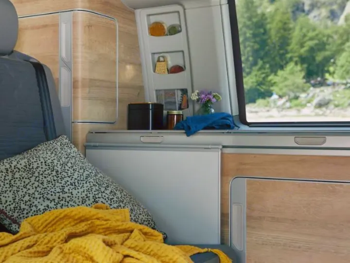 Compact RV rental interior featuring a cozy seating area, integrated fridge, and kitchen with a scenic nature view.. Volkswagen California Coast T6.1 DSG7 2023
