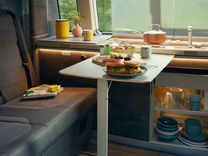 Interior of a modern RV rental, showing a cozy dining setup with sandwiches & fruit, plus a compact kitchen & seating.. Volkswagen California Coast T6.1 DSG7 2023