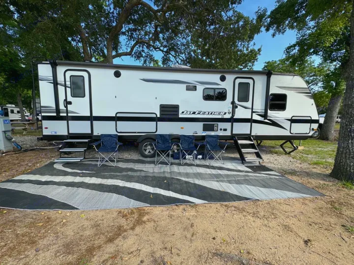 An RV rental, a Jay Feather travel trailer, is set up with an outdoor mat and chairs at a sunny campsite.. Jayco Jay Feather 2022