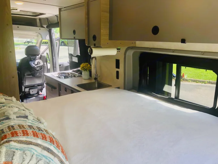 RV rental interior: comfy bed, compact kitchen with sink/stove, and cabinets. Open door reveals park view.. Winnebago Solis 2023