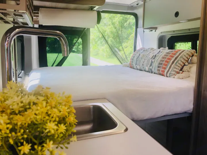 Cozy rv rental interior: bed with pillow, kitchen sink, and bright green outdoor view from rear windows.. Winnebago Solis 2023