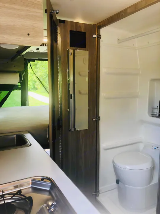 RV rental's compact interior: kitchen with stove, bed, and bathroom with shower and toilet. Scenic nature view from the rear.. Winnebago Solis 2023
