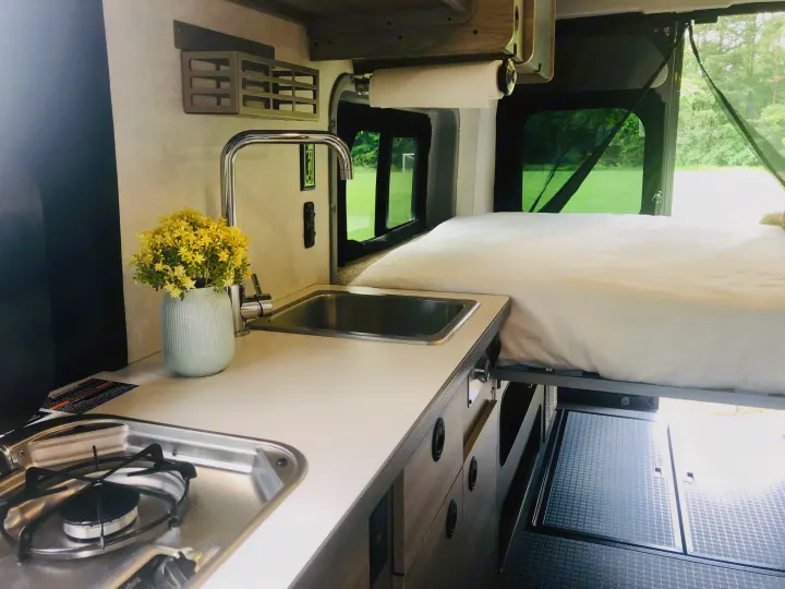 Interior of an RV rental showing a compact kitchen with a sink, faucet, stove, and an elevated bed.. Winnebago Solis 2023