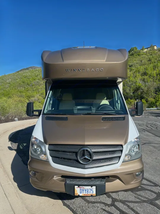 Front view of a brown and white Winnebago rv rental (Mercedes-Benz chassis) parked with green hills under a clear blue sky.. Winnebago View 2017