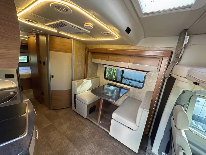 Modern RV rental interior showing a kitchen counter with sink, a dining booth and table, and overhead storage. The ceiling has strip lighting and vents.. Winnebago View 2017