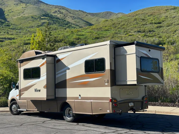 This beige and brown Winnebago View rv rental is parked with slide-outs extended, set against lush green mountains.. Winnebago View 2017