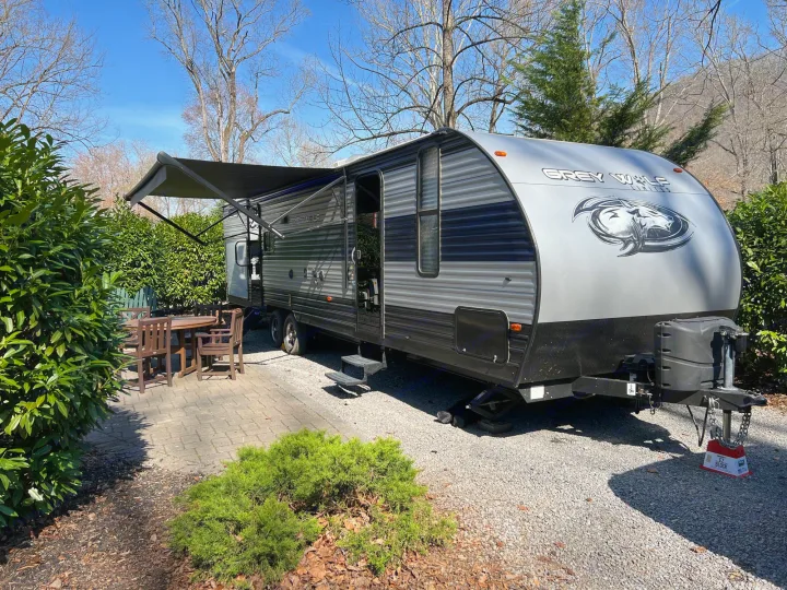 A Grey Wolf travel trailer, perfect for an rv rental, is set up with its awning extended at a sunny campsite with a patio.. Forest River Cherokee Grey Wolf 2021