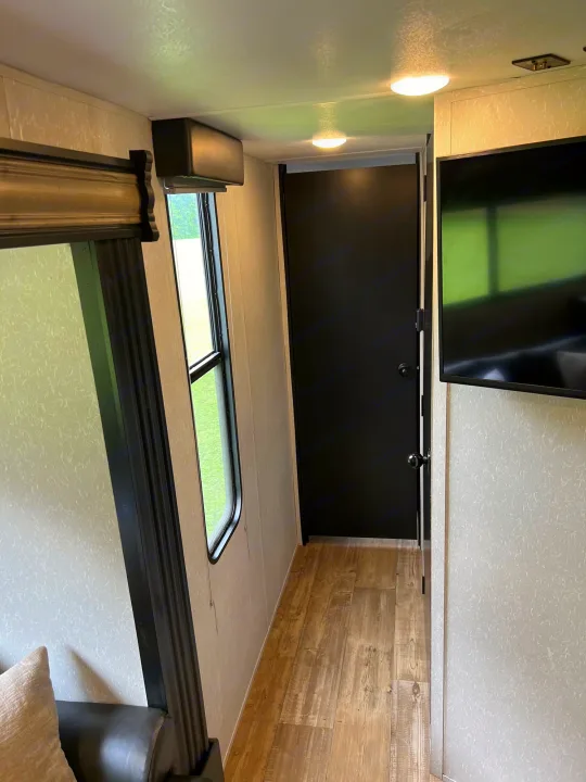 Inside an rv rental: a compact hallway with a black door, window showing green outside, and a wall-mounted TV.. Forest River Cherokee Grey Wolf 2021