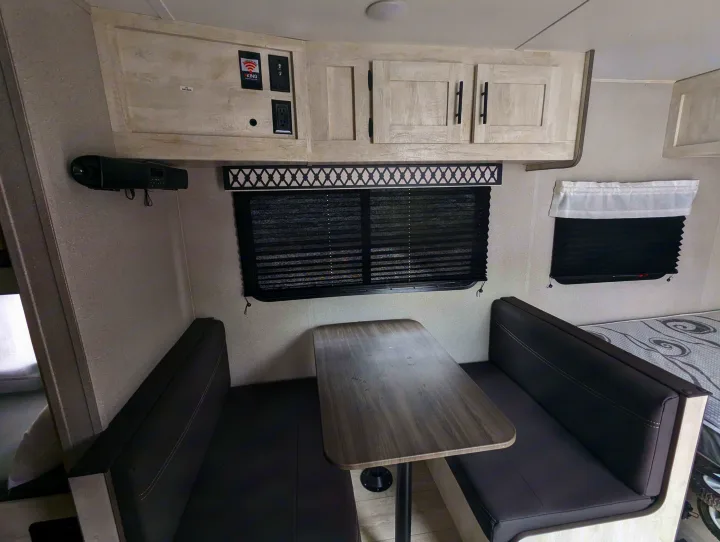 RV rental interior featuring a cozy dinette with a table, benches, overhead cabinets, window, and a bed.. Grand River Comfortline 20bh 2022