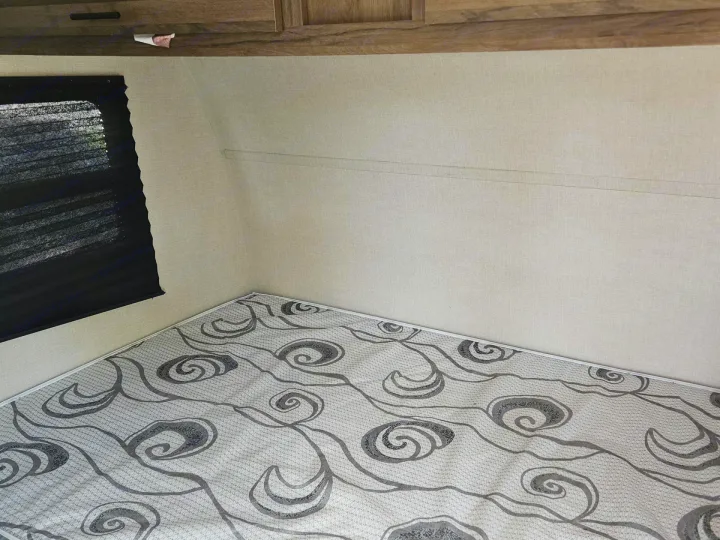 RV rental interior: light-walled sleeping area with a patterned mattress, black window blind, and wood cabinets overhead.. Avenger LT 2021