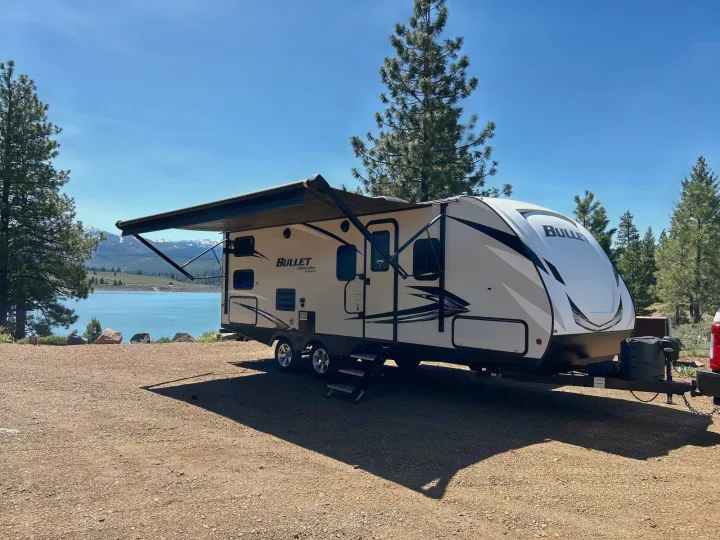Scenic RV rental: A travel trailer with an extended awning by a clear blue lake, snow-capped mountains, and pine trees.. Keystone RV Bullet Ultra Lite 2021