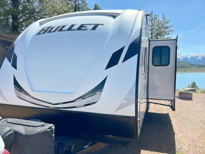 Explore with this white Bullet RV rental, parked near a blue lake and snowy mountains. Ideal for scenic getaways.. Keystone RV Bullet Ultra Lite 2021