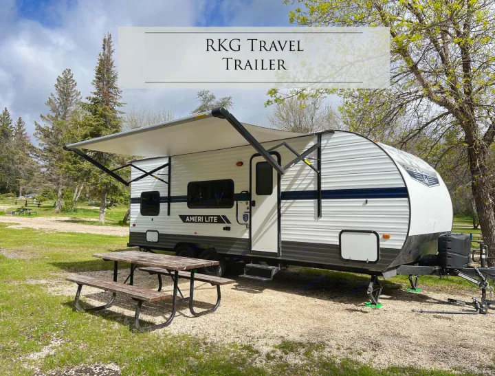 A white RKG Travel Trailer rv rental parked on a gravel campsite with an extended awning, picnic table, and trees.. Gulf Stream Ameri-Lite 2022