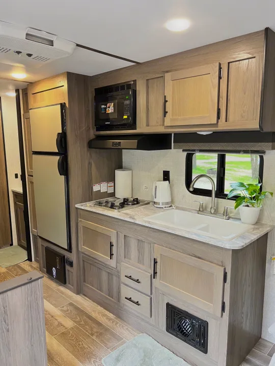 Explore this RV rental's modern kitchen: light wood cabinetry, stove, sink, microwave, and fridge for comfortable travel.. Gulf Stream Ameri-Lite 2022