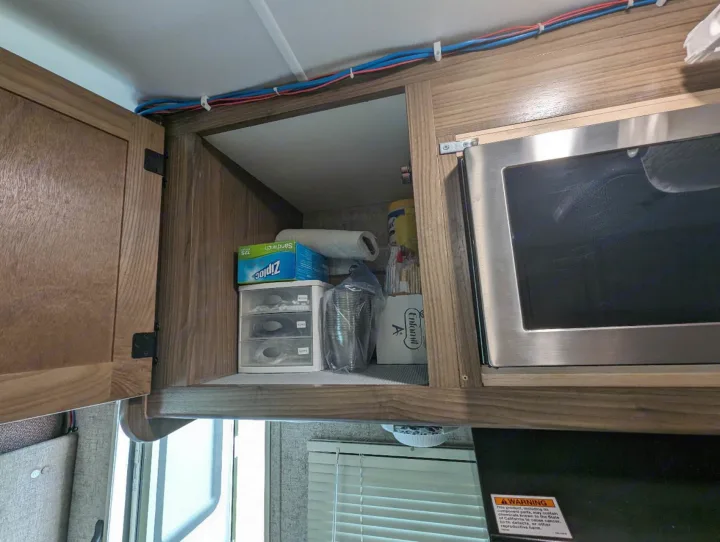 An RV rental's kitchen cabinet is open, revealing stored Ziploc bags, paper towels, and cups beside a microwave.. Forest River Coachmen Freelander 2018