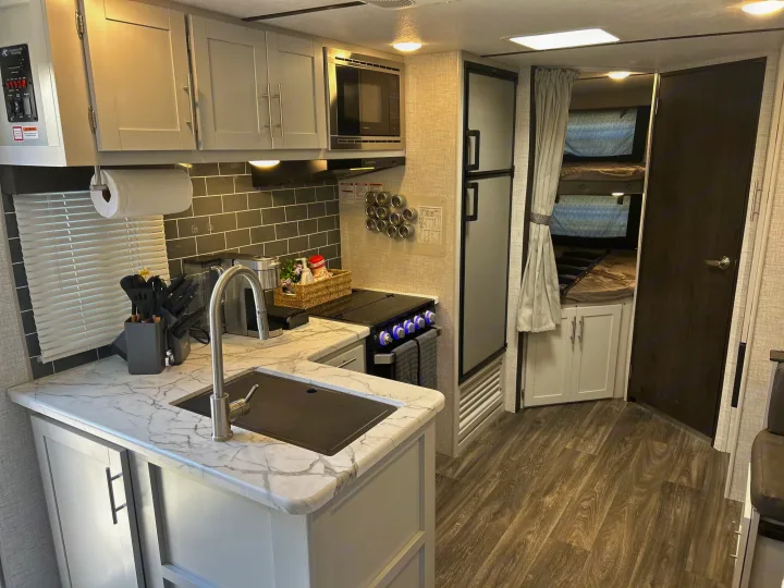 Modern rv rental kitchen with white cabinets, marble-look counters, stove, microwave, and visible bunk beds.. Keystone RV Bullet Ultra Lite 2021