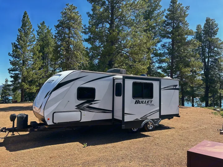 RV rental parked at a scenic lakeside campsite under a clear blue sky, surrounded by tall pine trees.. Keystone RV Bullet Ultra Lite 2021