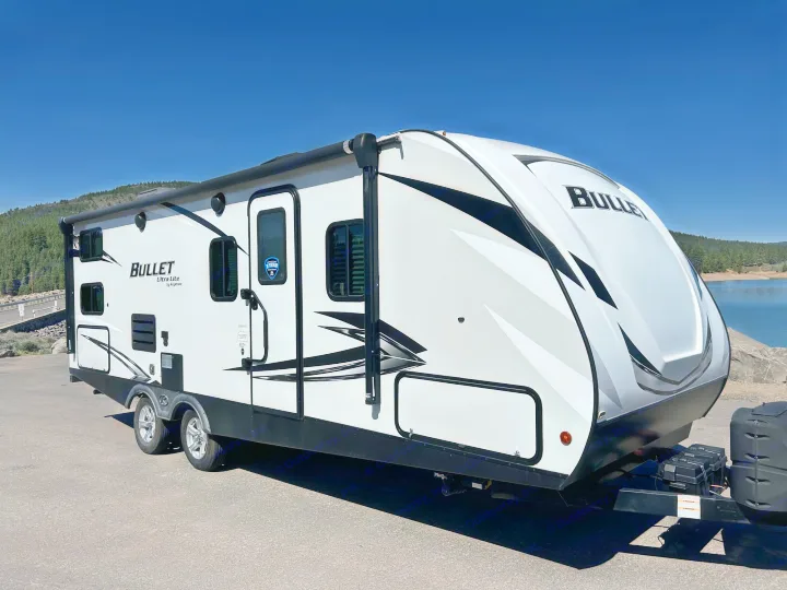 This white Keystone Bullet travel trailer, ideal for an RV rental, is parked by a scenic lake on a sunny day.. Keystone RV Bullet Ultra Lite 2021
