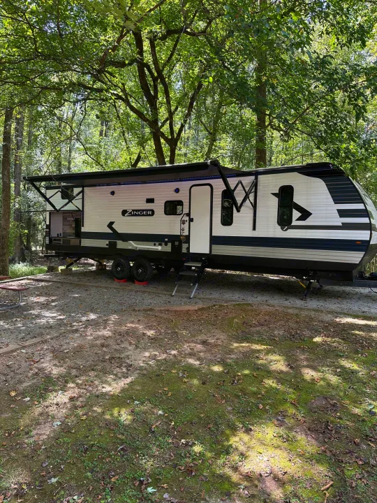 A Zinger travel trailer RV rental parked at a wooded campsite, featuring an extended awning and open outdoor kitchen.. Crossroads RV Zinger 2022