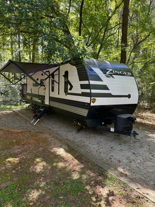 Ready for adventure: a Zinger travel trailer RV rental set up with its awning extended on a shaded gravel campsite among trees.. Crossroads RV Zinger 2022