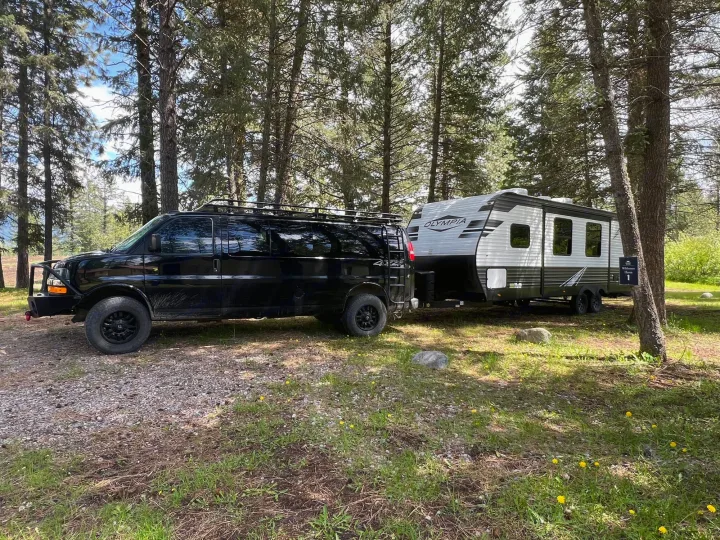 Black van towing an Olympia RV rental at a peaceful wilderness campsite, surrounded by tall trees.. Highland Ridge Open Range Olympia 2022