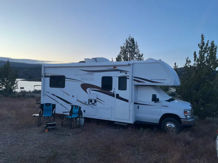 Enjoying the outdoors with an RV rental! A white motorhome parked at a tranquil lakeside campsite with chairs and a grill.. Jamboree Searcher 2013